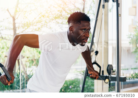 Muscular African American sportsman doing cable fly with exercise machine standing against window during training in gym Muscular African American sportsman doing cable fly with exercise machine standing against window during training in gym 67193010