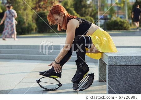 Beautiful woman in sportswear jumping in a kangoo jumps shoes at the street on summer's sunny day Beautiful woman in sportswear jumping in a kangoo jumps shoes at the street on summer's sunny day 67220993