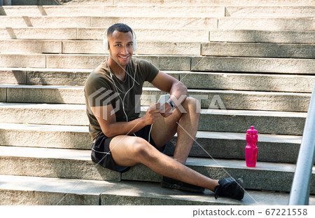 Happy black sportsman with headphones and mobile phone sitting on stairs and listening to music after workout 67221558