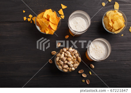 Nachos, chips, pistachios in glass bowls and scattered on wooden table, near are beer with foam in glasses 67221847