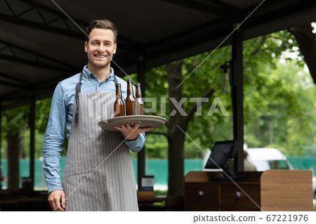 Work a waiter. Smiling handsome man in apron holds tray with bottles of beer in hands Work a waiter. Smiling handsome man in apron holds tray with bottles of beer in hands 67221976