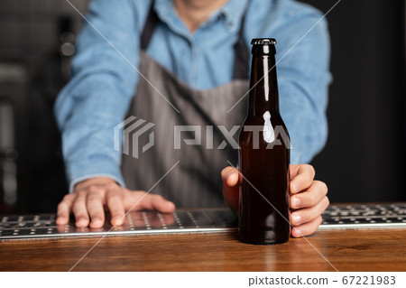 Barman in apron rests on wooden bar counter and serves bottle of beer without label 67221983