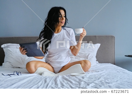 Beautiful woman sitting in bed with cup of coffee and tablet 67223671