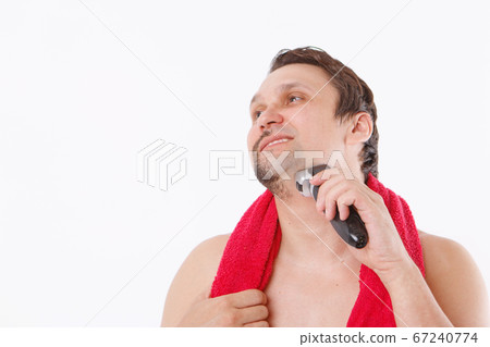 isolated on a white background: a man shaves his stubble. the guy cleans his beard with an electric razor. morning treatments in the bathroom. red towel around her neck. copy space 67240774