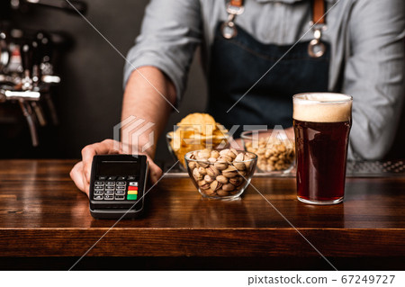 Modern payment at bar. Barman in apron gives to customer a glass of dark beer, snacks and terminal Modern payment at bar. Barman in apron gives to customer a glass of dark beer, snacks and terminal 67249727