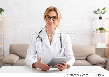 Contact with patients online. Woman doctor with glasses and white coat holds digital tablet in hands 67249870