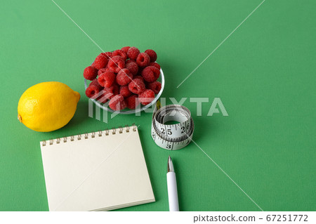 Inch measuring tape roll, lemon, raspberries, paper notepad and white pen on a green background. Inch measuring tape roll, lemon, raspberries, paper notepad and white pen on a green background. 67251772