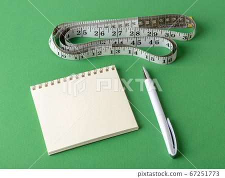 Measuring tape with centimeters and inches, paper notepad and white pen on a green background. 67251773