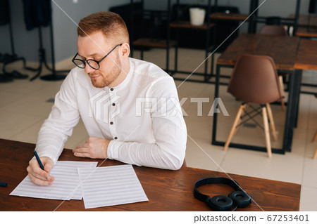 Talented young composer wearing stylish eyeglasses writing musical notes with pen on sheet music Talented young composer wearing stylish eyeglasses writing musical notes with pen on sheet music 67253401