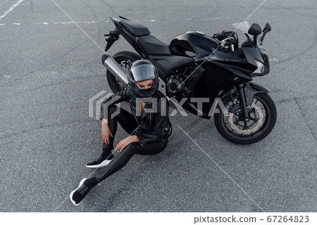 Biker woman in black leather jacket and full face helmet sits near stylish sports motorcycle at urban parking. Traveling and active lifestyle concept. 67264823