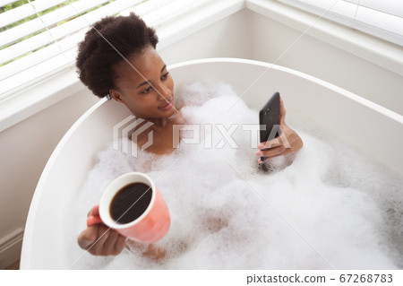 Woman with coffee cup using smartphone in a bathtub Woman with coffee cup using smartphone in a bathtub 67268783