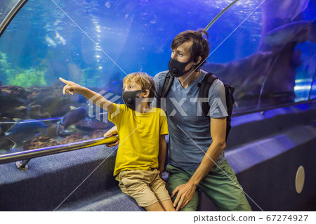 Dad and son in medical masks look at the fish in the aquarium in oceanarium. People walk in medical masks after the end of the coronovirus 67274927