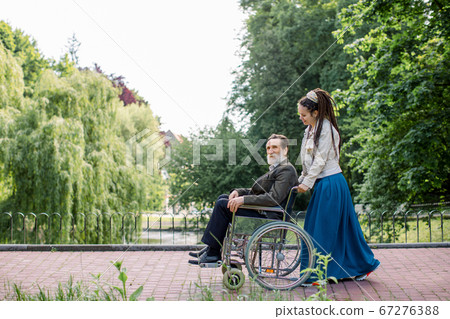 Side view of young modern hipster woman with long dreadlocks, assisting her disabled grandfather on wheelchair in park. Happy elderly man in wheelchair with young girl social worker in the park. Side view of young modern hipster woman with long dreadlocks, assisting her disabled grandfather on wheelchair in park. Happy elderly man in wheelchair with young girl social worker in the park. 67276388
