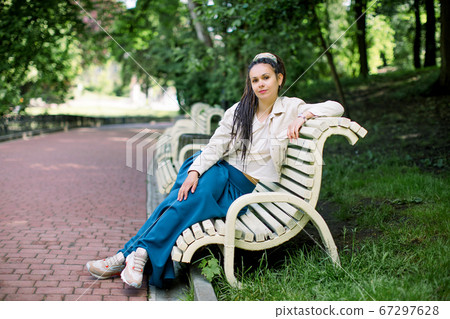 Beautiful girl with dreadlocks, in a trendy long blue skirt and white tshirt, sitting on a bench in a park on the trees and alley background 67297628