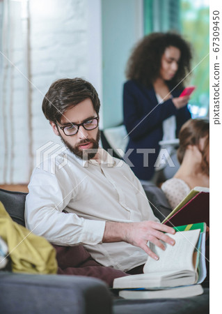 Bearded guy with glasses sitting on sofa with books, two girls 67309450