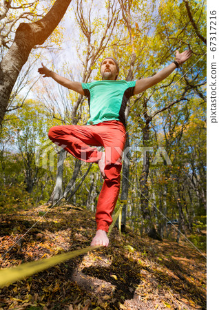 A bearded man aged walks along a slackline in the autumn forest on a sunny afternoon. The concept of Leisure and Recreation of active middle-aged people 67317216