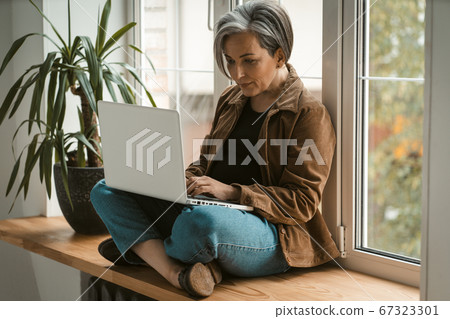 Serious mature woman in casual uses laptop computer sitting crossed legs on wooden window sill. Toned image. Freelance concept 67323301