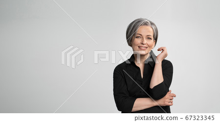 Mature Caucasian woman smiling charmingly with hand near face. Graying elegant woman with short bob hairstyle looks at camera posing on white background. Blank or template with text space at left 67323345