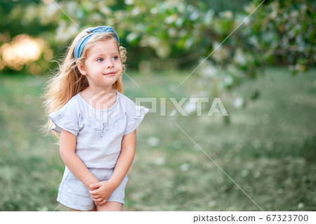 A charming little girl with long blond hair playing against the background of nature. A happy child enjoys summer time in the Park. 67323700