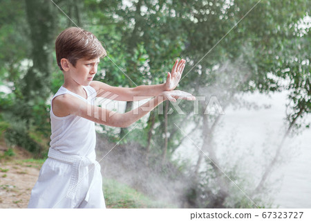 One little boy in a white kimono during a karate kata training exercise in the summer outdoors. One little boy in a white kimono during a karate kata training exercise in the summer outdoors. 67323727