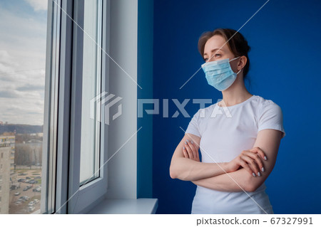 Pensive woman wearing medical face mask and looking out of window in blue room Pensive woman wearing medical face mask and looking out of window in blue room 67327991