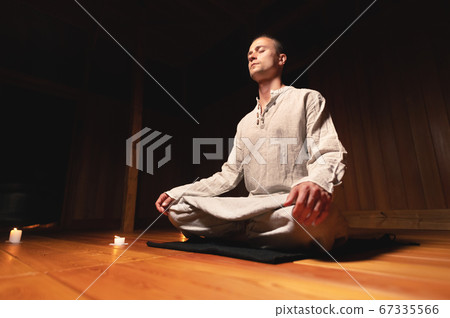 A portrait of an attractive Caucasian man sits in cotton robes in a lotus pose in a dark wooden practice room surrounded by candles A portrait of an attractive Caucasian man sits in cotton robes in a lotus pose in a dark wooden practice room surrounded by candles 67335566