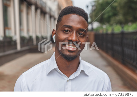 Portrait of a smiling black businessman with wireless earphones walking outdoors 67339873