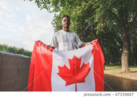Handsome Afro American man with Canadian flag seriously looking at camera, standing outdoors. 67339887