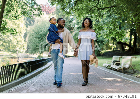 Full length portrait of adorable african family of three, father, mother and child, walking in the park. Handsome father holds his daughter in arms and holds hand of his charming wife 67348191