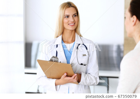 Doctor woman at work in hospital excited and happy of consulting female patient. Blonde physician controls medication history records and exam results while using clipboard. Medicine and healthcare 67350079