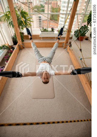Young sportsman in activewear hanging over the floor during aerial yoga workout 67358806