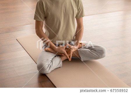 Low section of young man sitting on mat and keeping hands between crossed legs 67358932