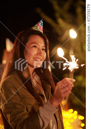 Happy young Asian woman in birthday cap holding bengal light at outdoor party 67359279