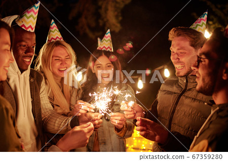 Cheerful intercultural friends in birthday caps holding bengal lights at party 67359280