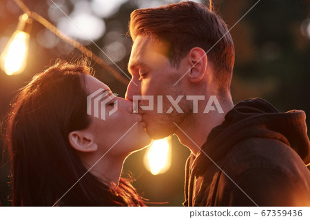 Happy young amorous couple kissing on summer evening against street lights Happy young amorous couple kissing on summer evening against street lights 67359436