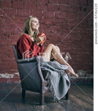 Young smiling woman holding red cup of coffee or tea drinking warm beverage sitting in cozy chair at loft apartment. Happy healthy lady relaxing with morning drink in hands enjoying peace of mind. Young smiling woman holding red cup of coffee or tea drinking warm beverage sitting in cozy chair at loft apartment. Happy healthy lady relaxing with morning drink in hands enjoying peace of mind. 67362229
