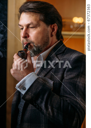 Reflective well-dressed mature bearded man in glasses smoking tobacco pipe 67372363