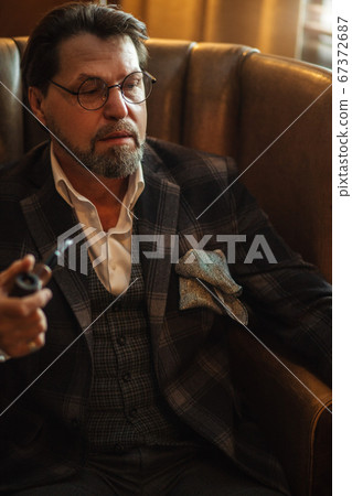 Reflective well-dressed mature bearded man in glasses smoking tobacco pipe Reflective well-dressed mature bearded man in glasses smoking tobacco pipe 67372687
