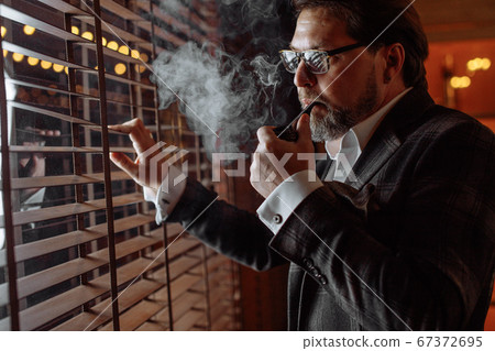 Reflective well-dressed mature bearded man in glasses smoking tobacco pipe Reflective well-dressed mature bearded man in glasses smoking tobacco pipe 67372695