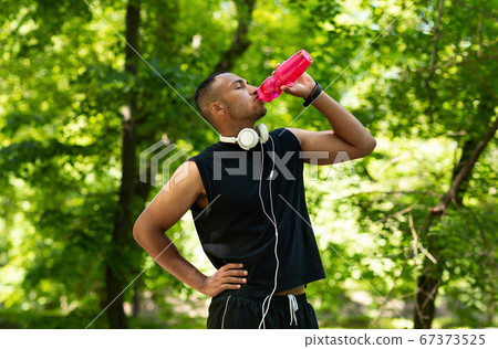 Thirsty young athlete drinking water from bottle after his training at park Thirsty young athlete drinking water from bottle after his training at park 67373525
