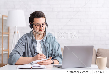 Studying and writing in notebook. Busy guy with glasses and headset sitting at table and look at laptop 67373710