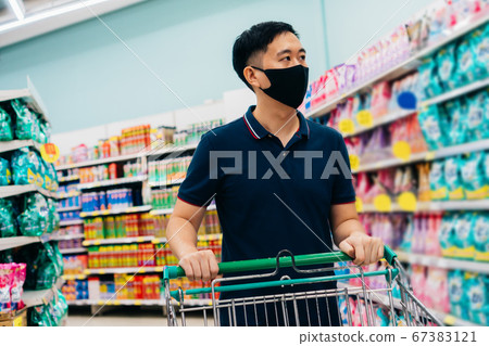 Young adult Asian man wearing a face mask while shopping with cart trolley in grocery supermarket store. Young adult Asian man wearing a face mask while shopping with cart trolley in grocery supermarket store. 67383121