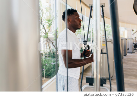 Muscular African American sportsman doing cable fly with exercise machine standing against window during training in gym Muscular African American sportsman doing cable fly with exercise machine standing against window during training in gym 67383126