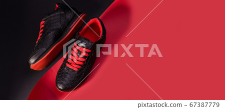 Fashionable snakeskin leather sneakers. Black sneakers with bright red bootlaces against a black and red background. Fashionable snakeskin leather sneakers. Black sneakers with bright red bootlaces against a black and red background. 67387779