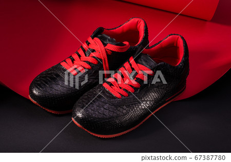 Fashionable snakeskin leather sneakers. Black sneakers with bright red bootlaces against a black and red background. Fashionable snakeskin leather sneakers. Black sneakers with bright red bootlaces against a black and red background. 67387780