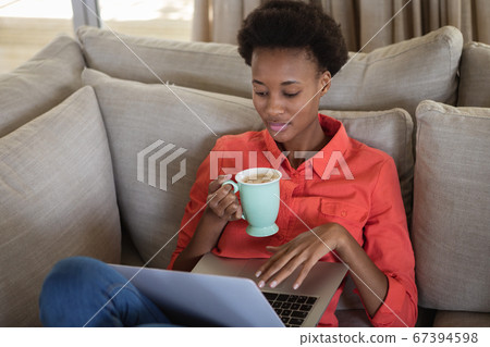 Woman with coffee cup using laptop while sitting on the couch Woman with coffee cup using laptop while sitting on the couch 67394598