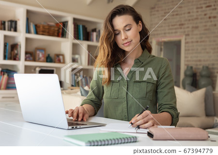 Woman taking notes while using laptop at home Woman taking notes while using laptop at home 67394779