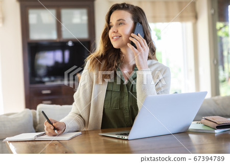 Woman taking notes while talking on smartphone at home Woman taking notes while talking on smartphone at home 67394789