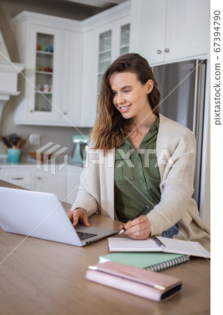 Woman taking notes while using laptop in the kitchen Woman taking notes while using laptop in the kitchen 67394790