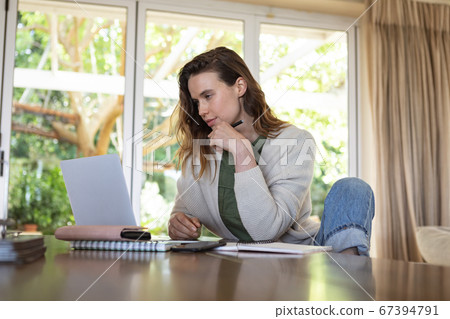 Woman taking notes while using laptop at home Woman taking notes while using laptop at home 67394791